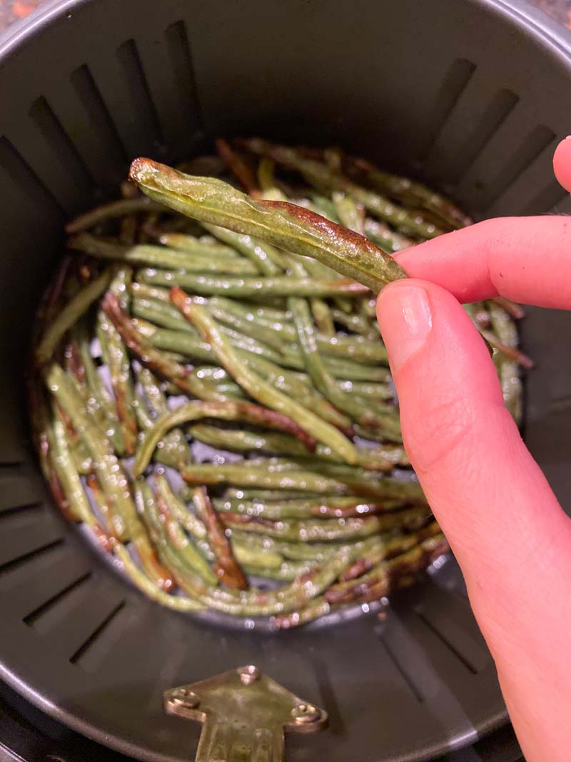 woman holding an air fryer green bean, with an air fryer basket full of them in the background