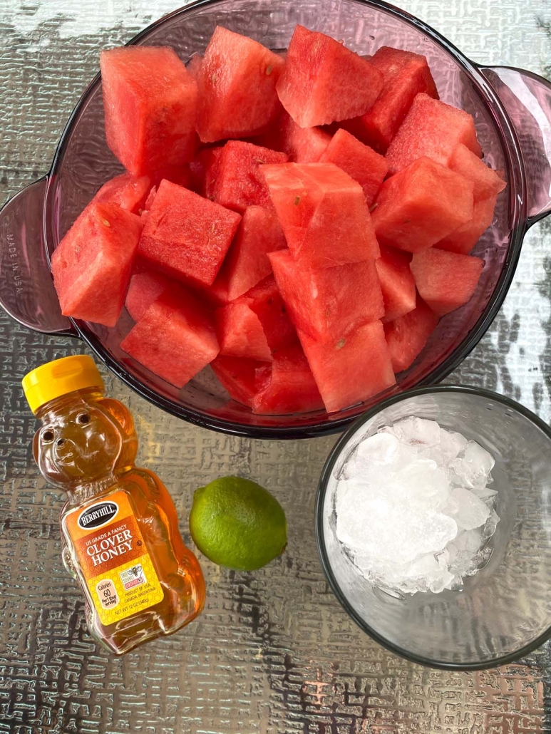 bowl of cut up watermelon next to honey, line, and glass of ice