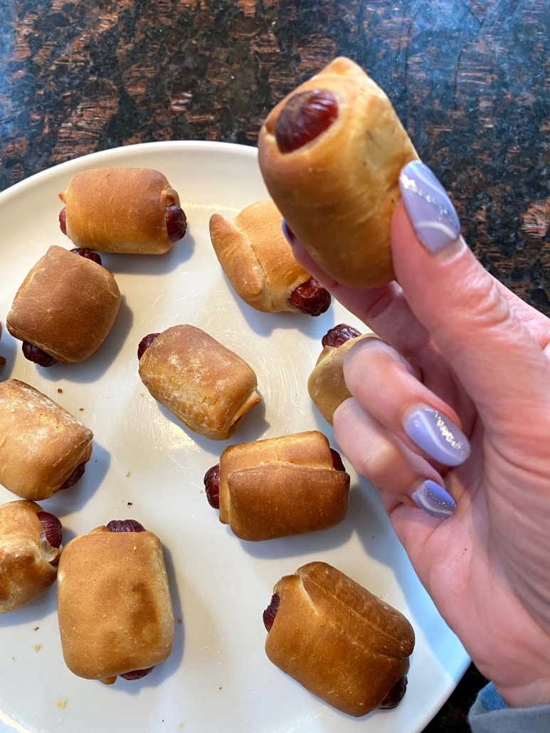hand holding mini bagel dog over plate with more bagel dogs