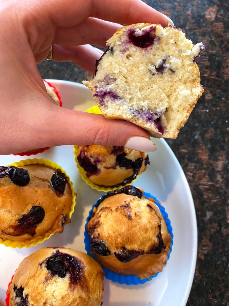 hand holding half a blueberry muffin above plate of muffins