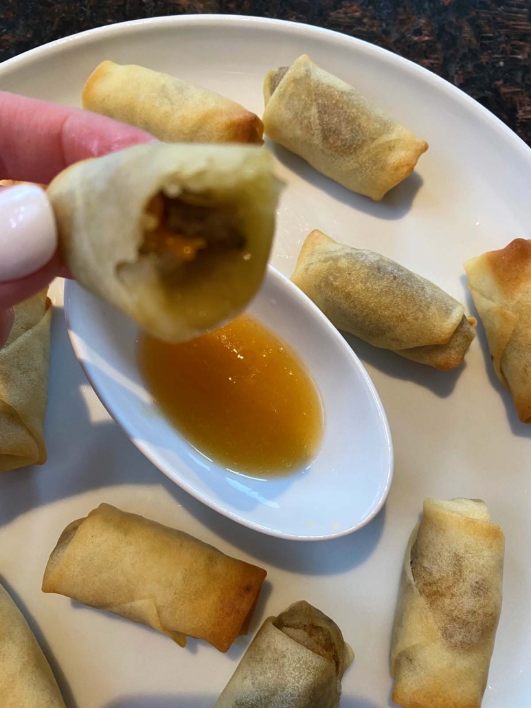 hand holding partially eaten spring roll above dipping sauce and plate of spring rolls