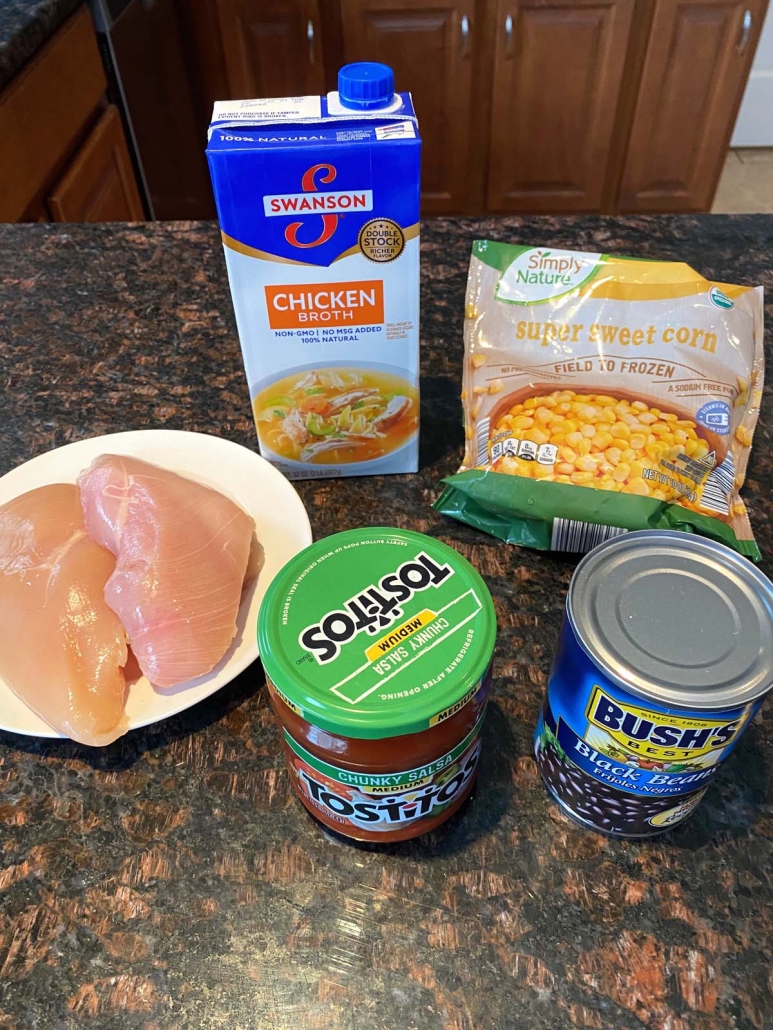 Chicken breast on plate, next to jar of salsa, chicken broth, can of beans, and bag of sweet corn