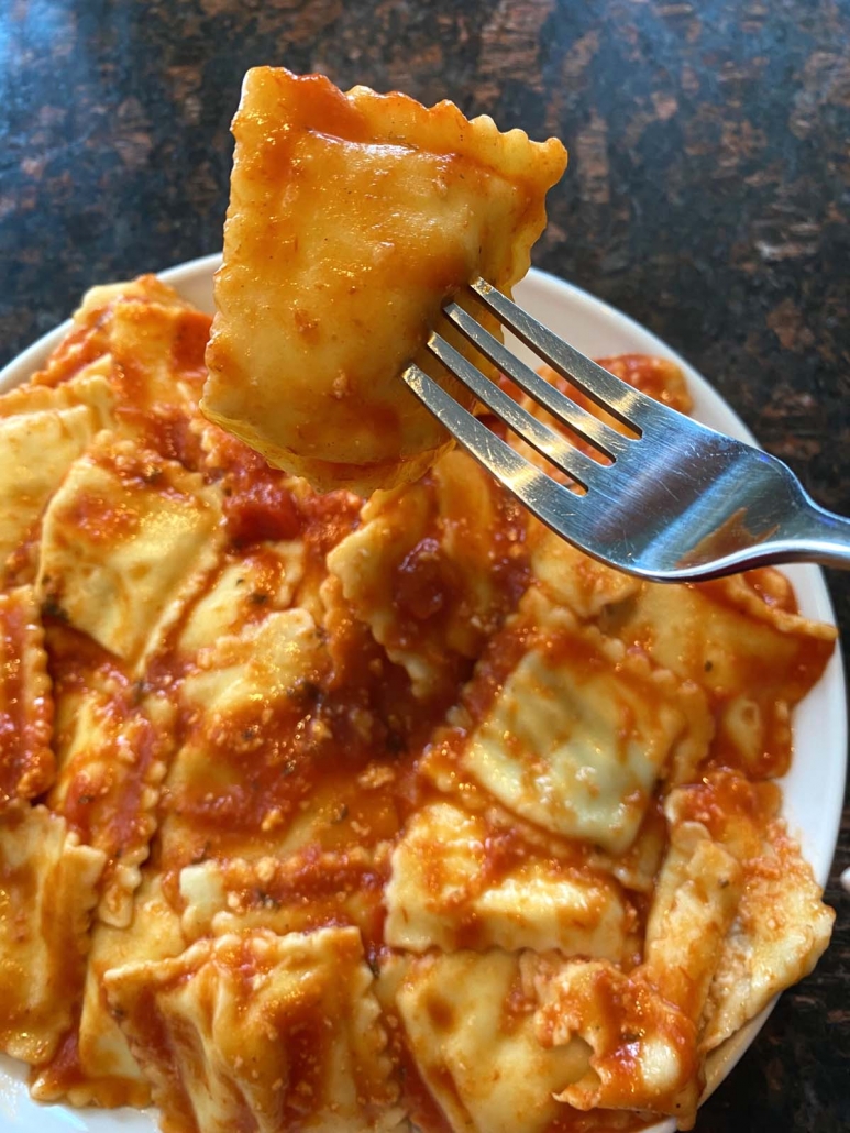 fork holding single ravioli above plate filled with ravioli and sauce