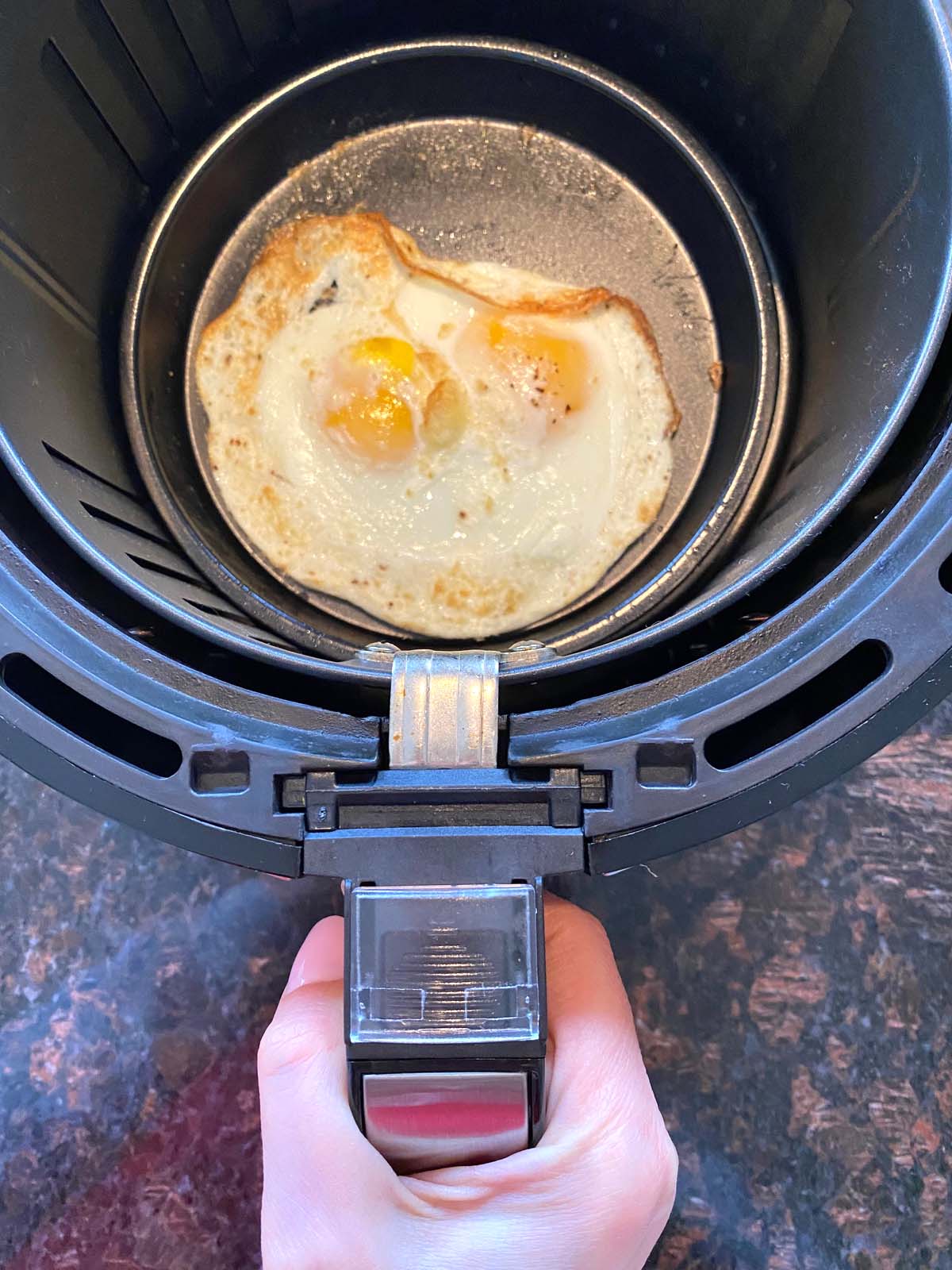 Hand holding handle of air fryer basket with 2 cooked eggs inside in a pan.