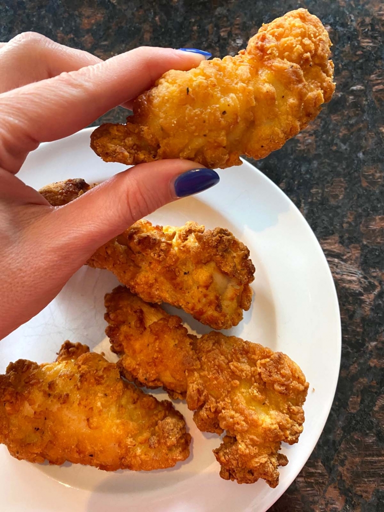 hand holding air fried chicken strip above a plate of more chicken strips