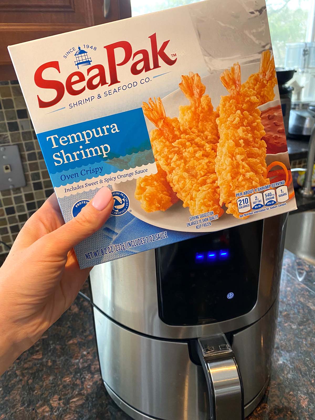 Hand holding a box of frozen SeaPak Tempura Shrimp infront of an air fryer.