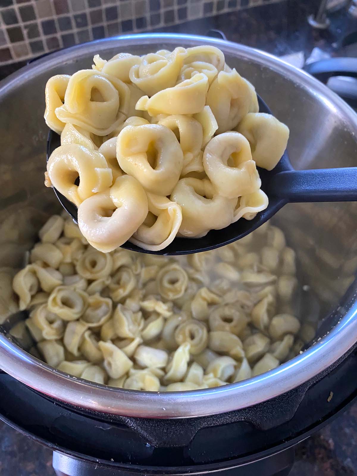 A spoon of cooked tortellini being held over an instant pot full of cooked tortellini.