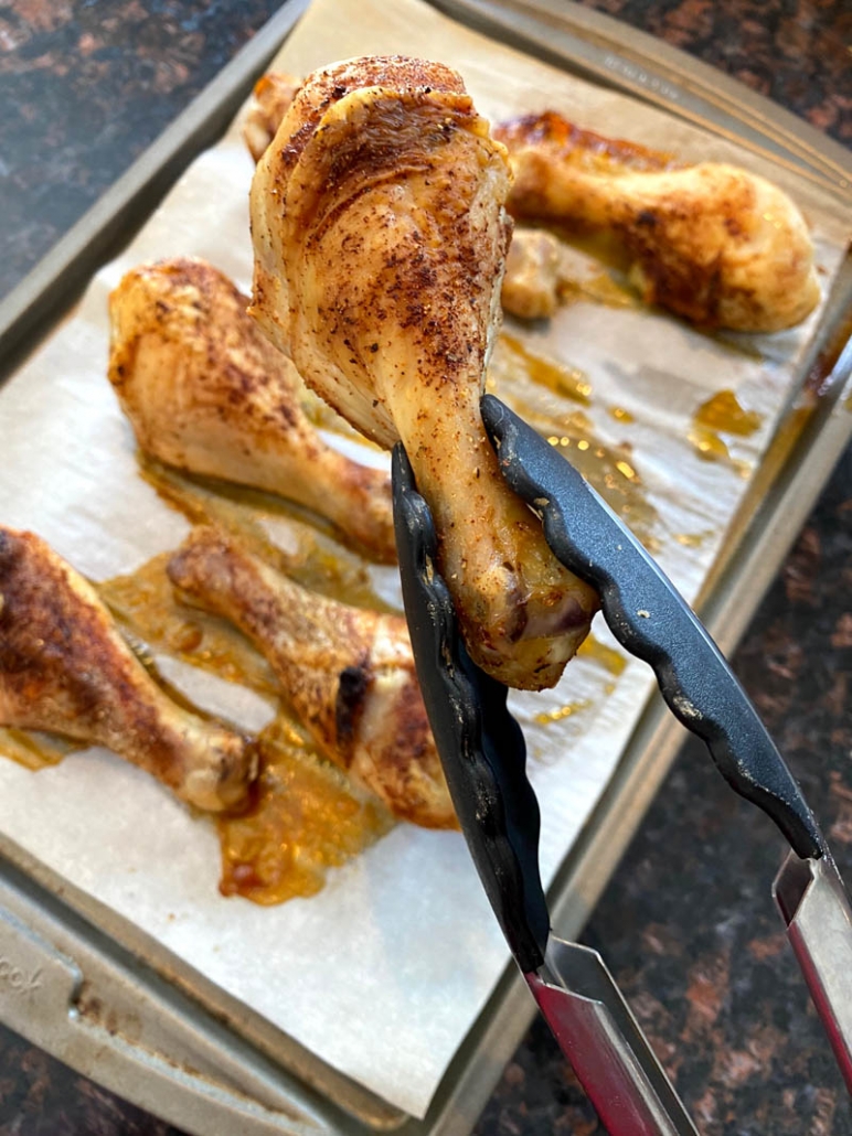 tongs holding oven baked drumstick above baking sheet of drumsticks