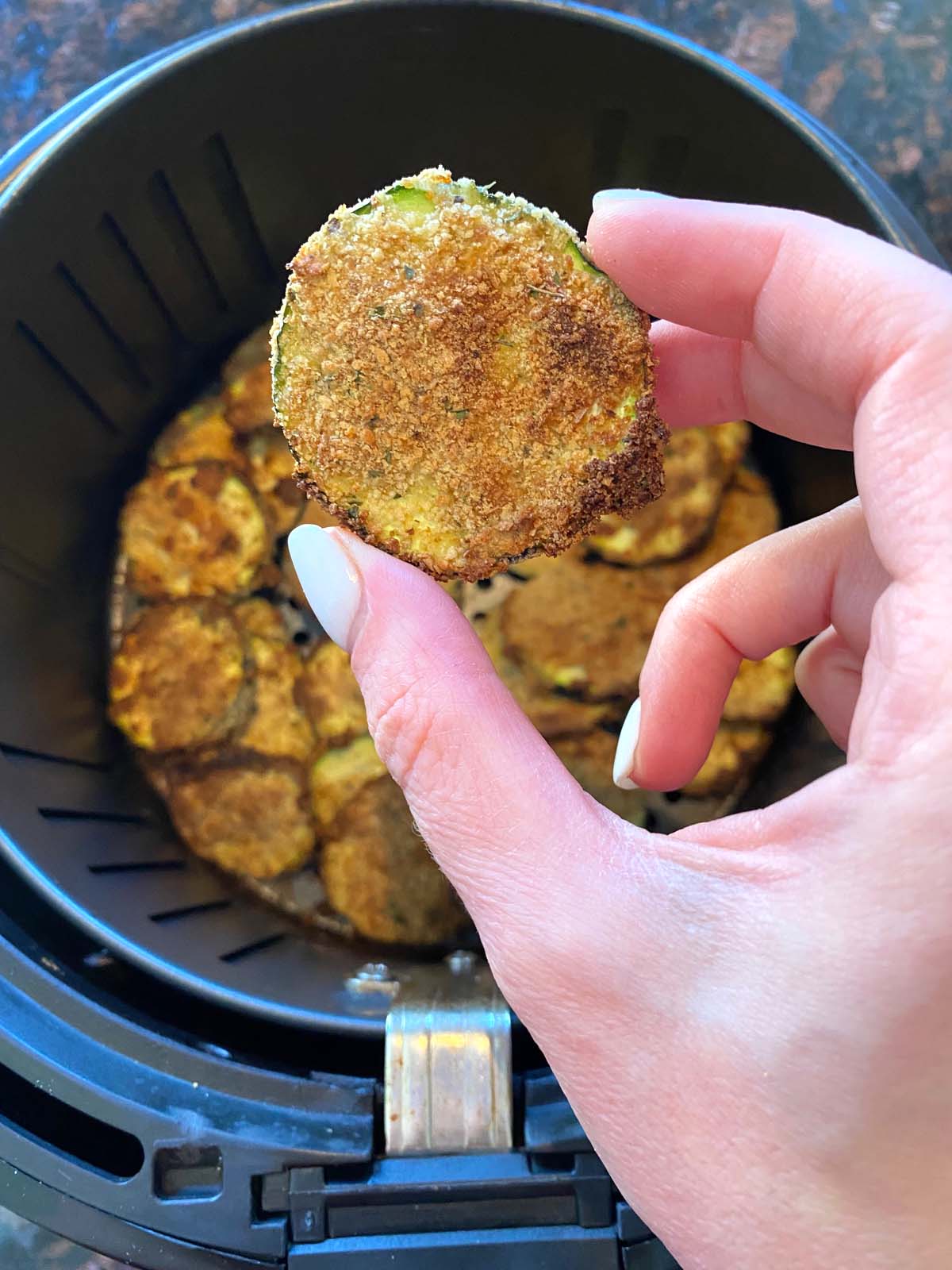 Cooked breaded zucchini in an air fryer with one piece being held up by hand.