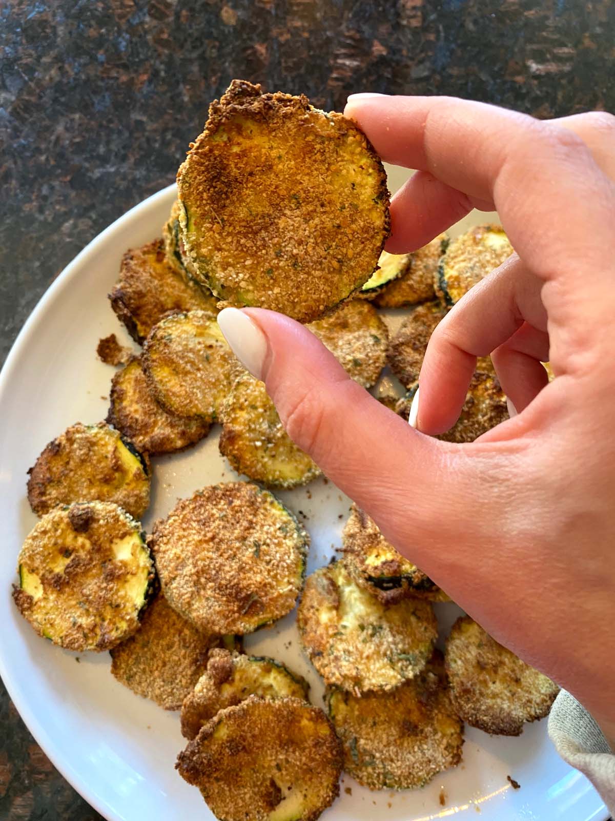 Breaded air fried zucchini chips on a white plate with one being held up by hand.