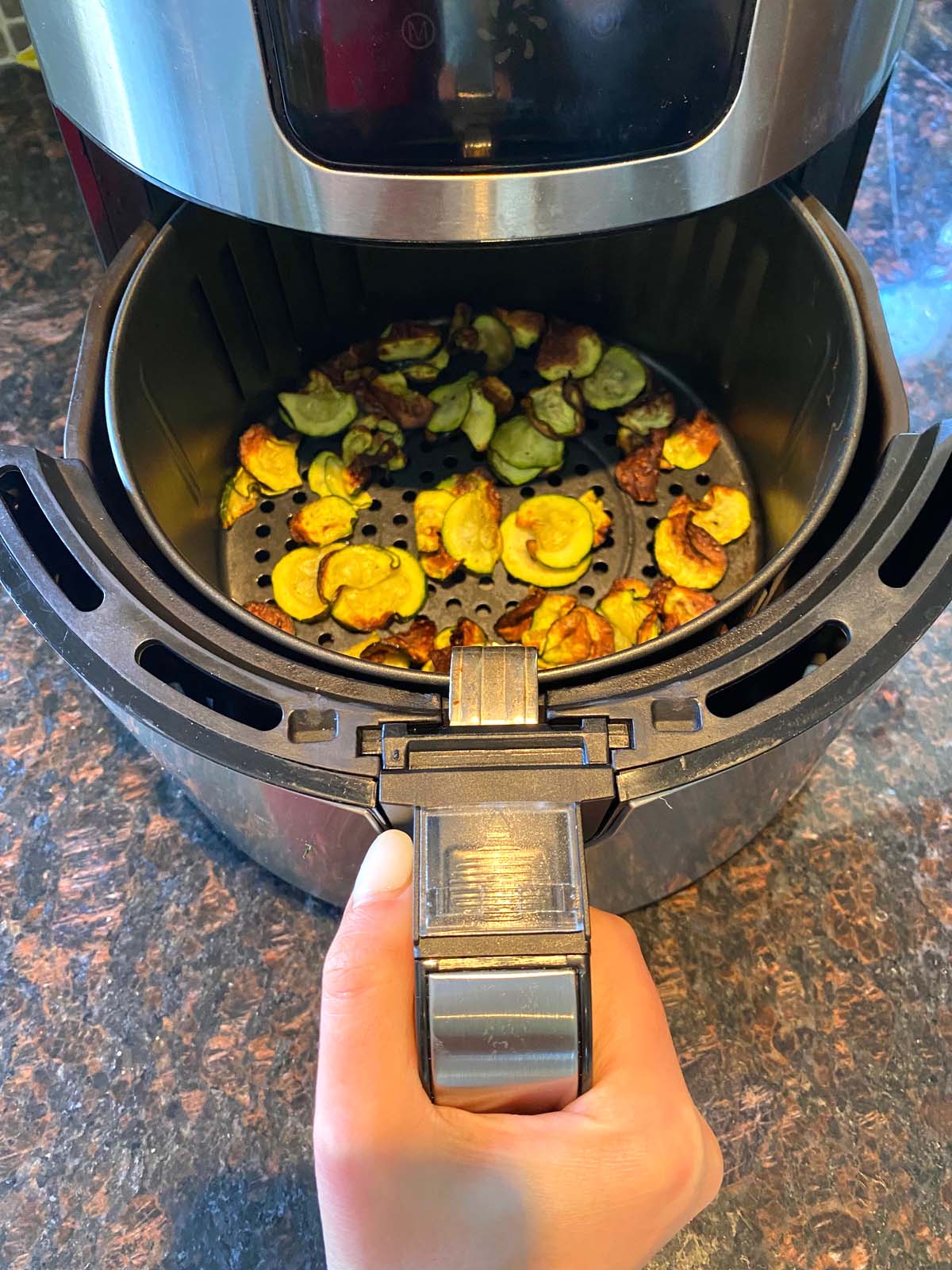 Hand pulling out basket from air fryer filled with zucchini chips.