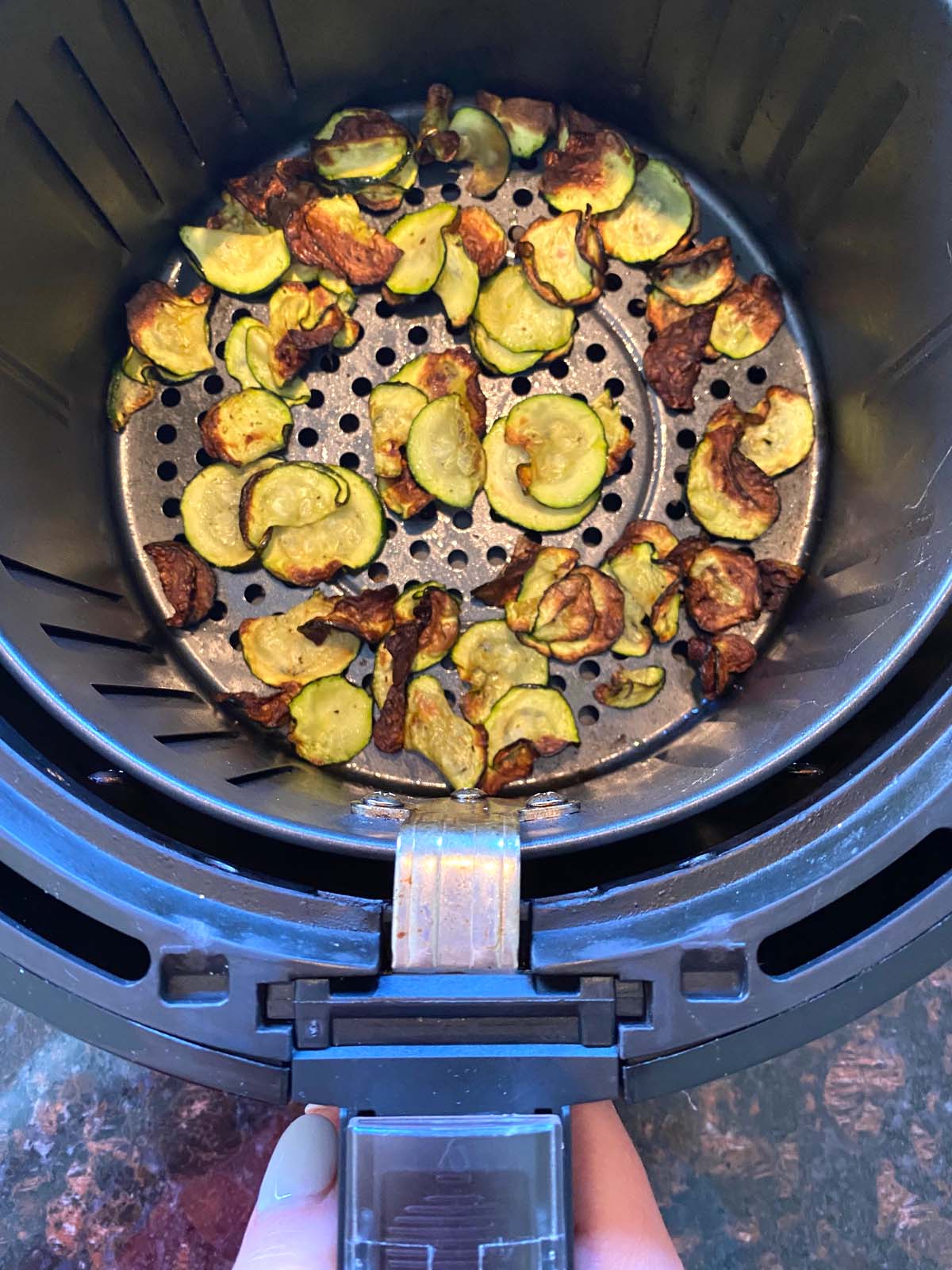 Hand pulling out basket from air fryer filled with zucchini chips.