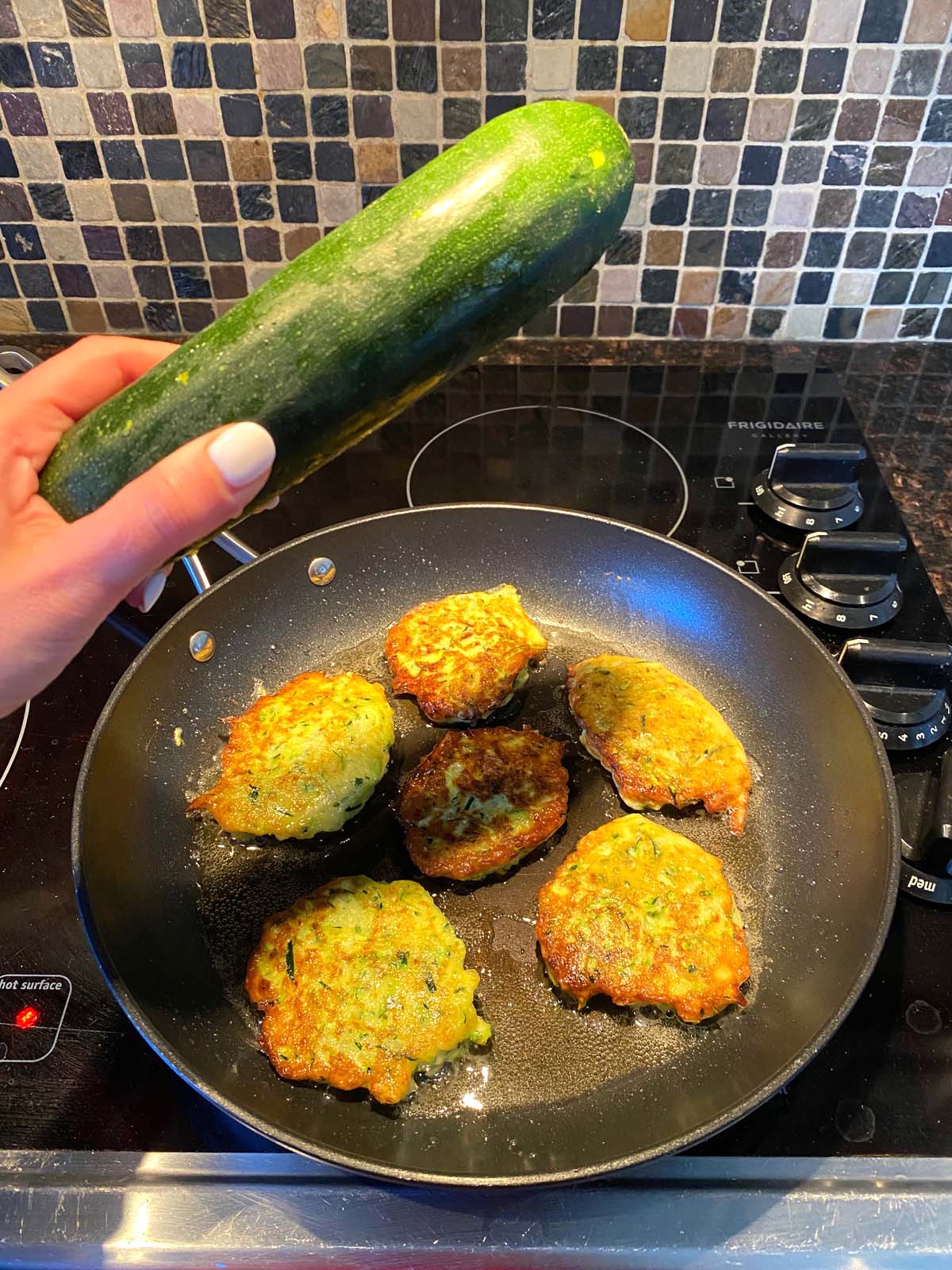 Zucchini pancakes in a frying pan with a raw zucchini being held up.