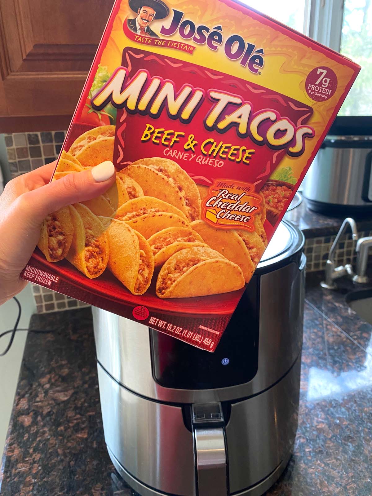 Hand holding a box of Jose Ole beef and cheese mini tacos in front of an air fryer.
