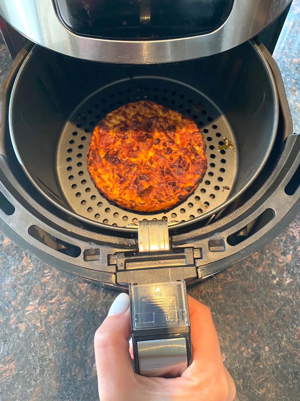 Single size deep dish frozen pizza in air fryer basket with hand inserting into machine.