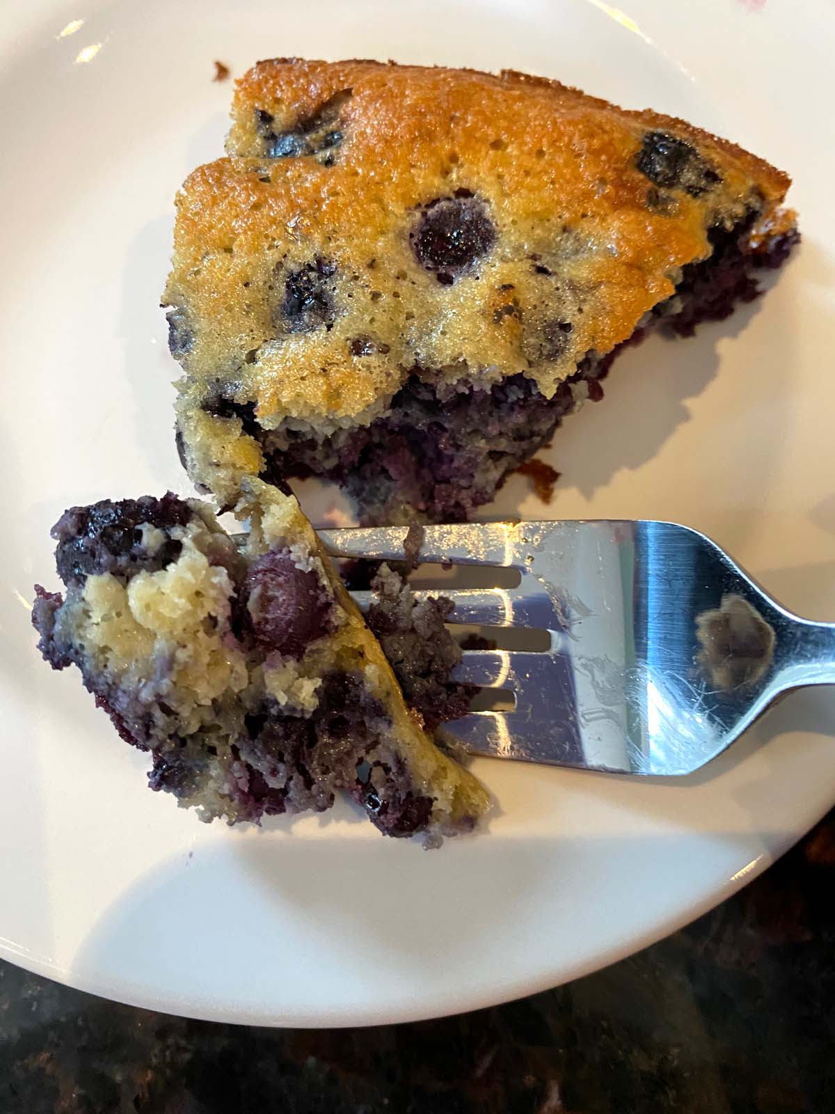 A slice of gluten free blueberry cake on a plate with a fork digging into it.