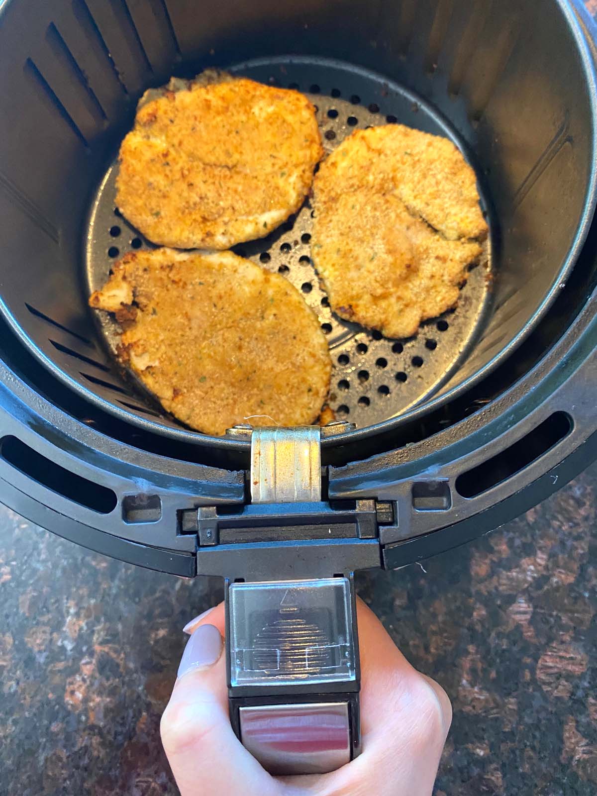 Handing pulling out basket from air fryer to show 3 pieces of turkey cutlets with crispy breading.