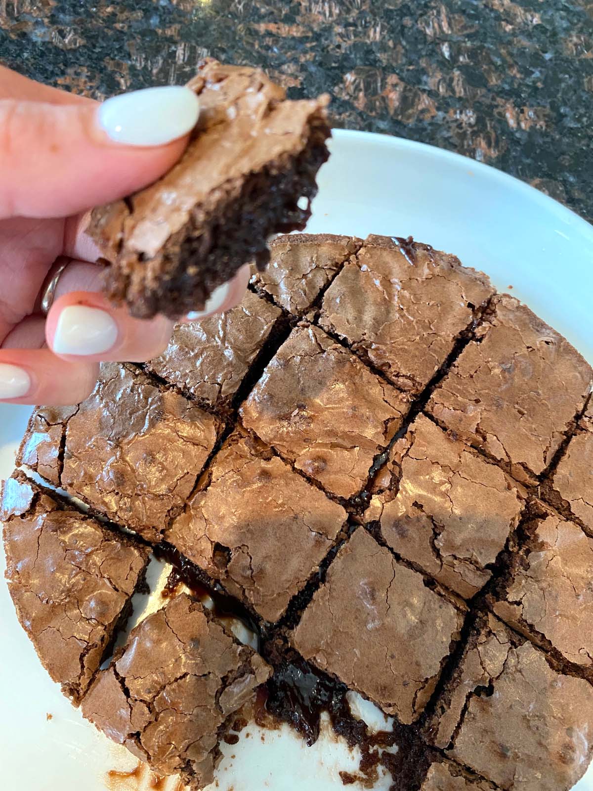 A hand holding up a piece of brownie with a plate of square cut brownies below.