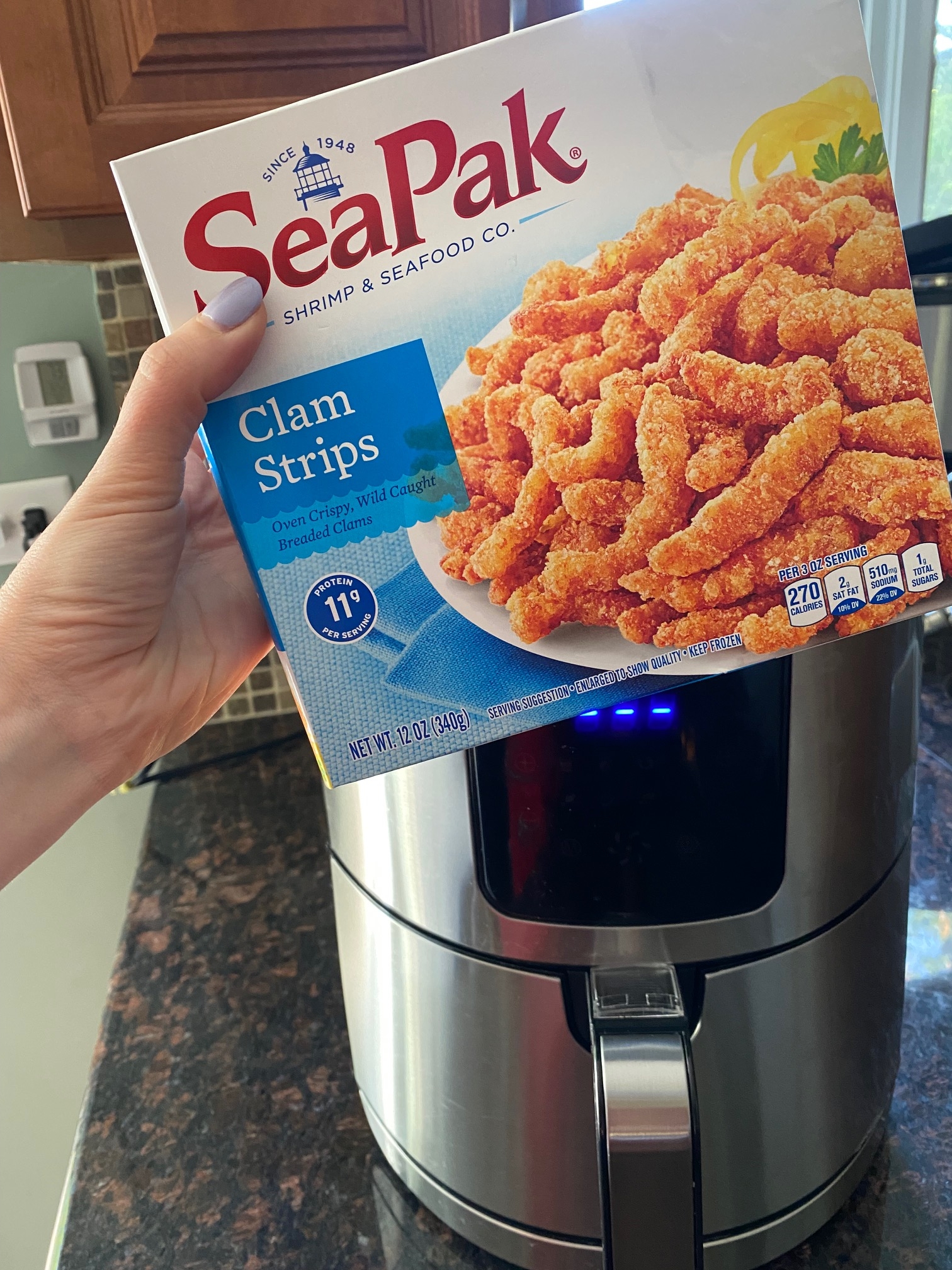 Hand holding box of SeaPak frozen clam strips in front of air fryer.