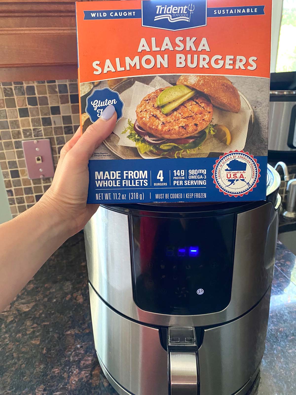 Box of frozen salmon burgers held in front an air fryer.