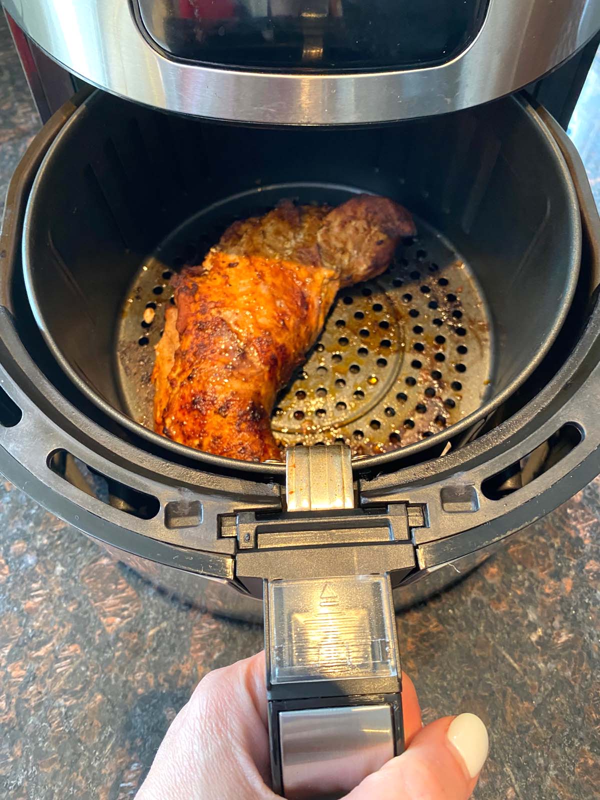 Hand removing basket from air fryer to show meat cooked with browning on top.