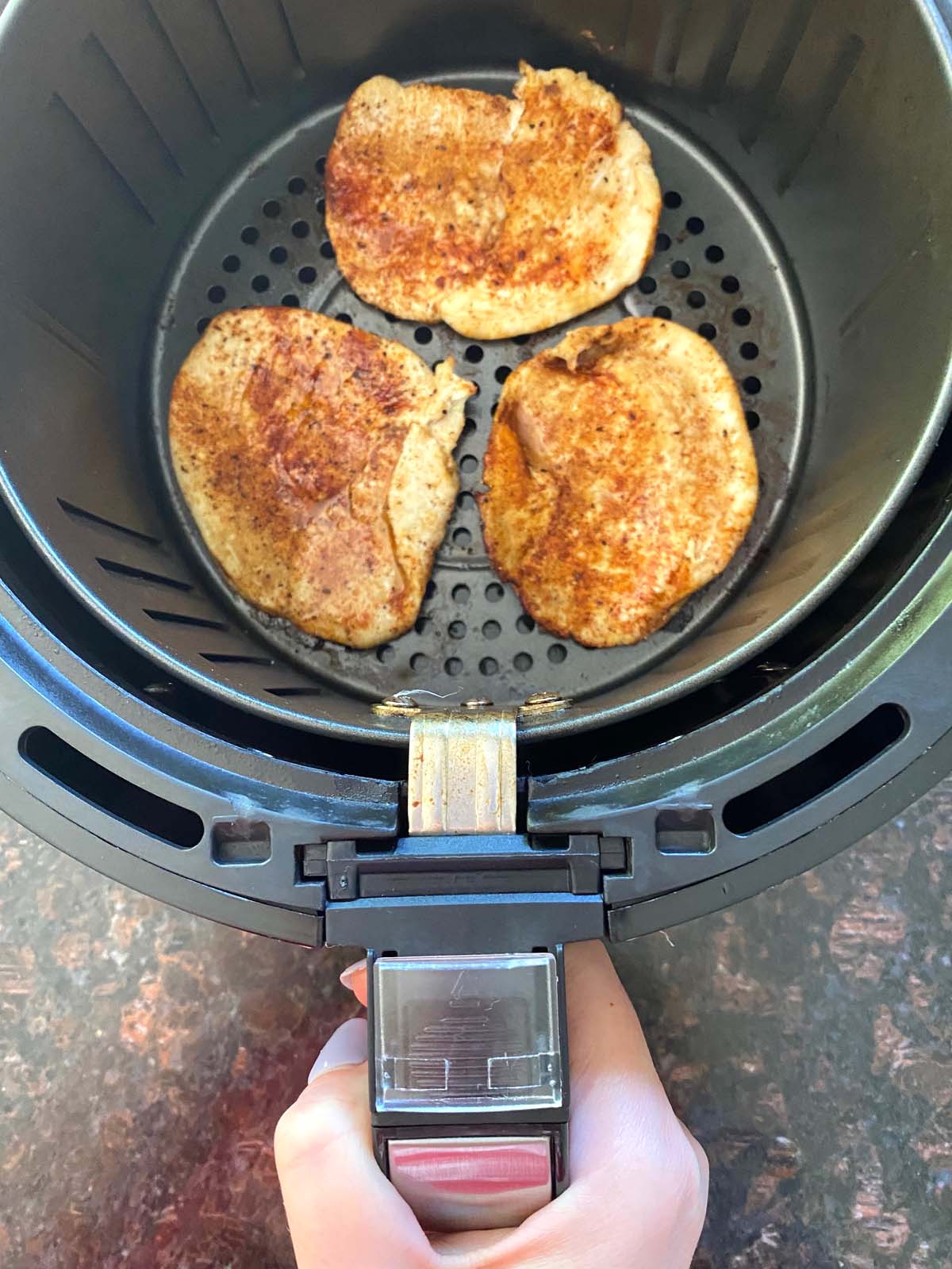 Hand holding handle of air fryer basket that has 3 pieces of cooked meat inside.