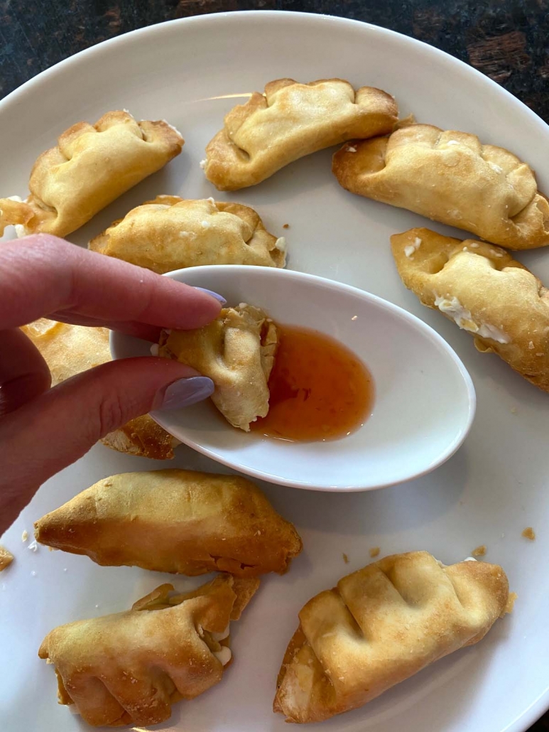 hand holding air fried cream cheese wonton in dipping sauce on plate with wontons