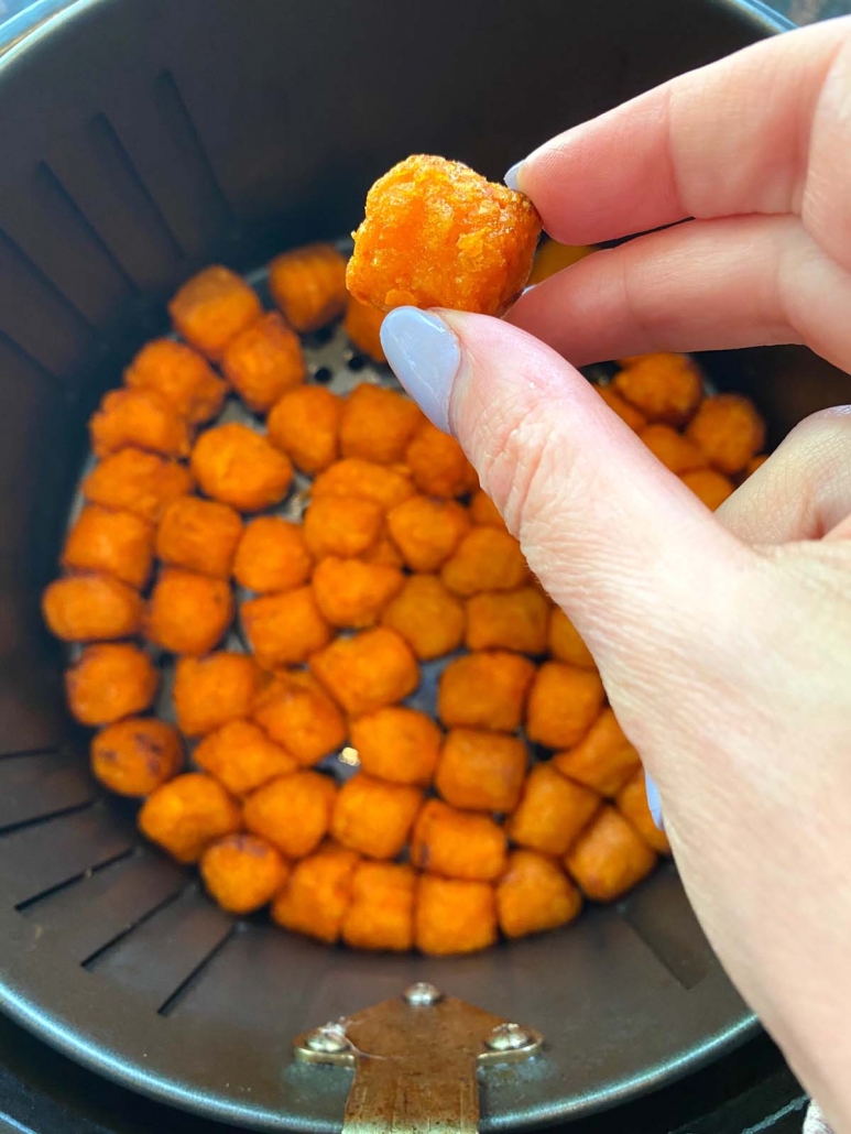 hand holding a cooked sweet potato puff above air fryer filled with more puffs