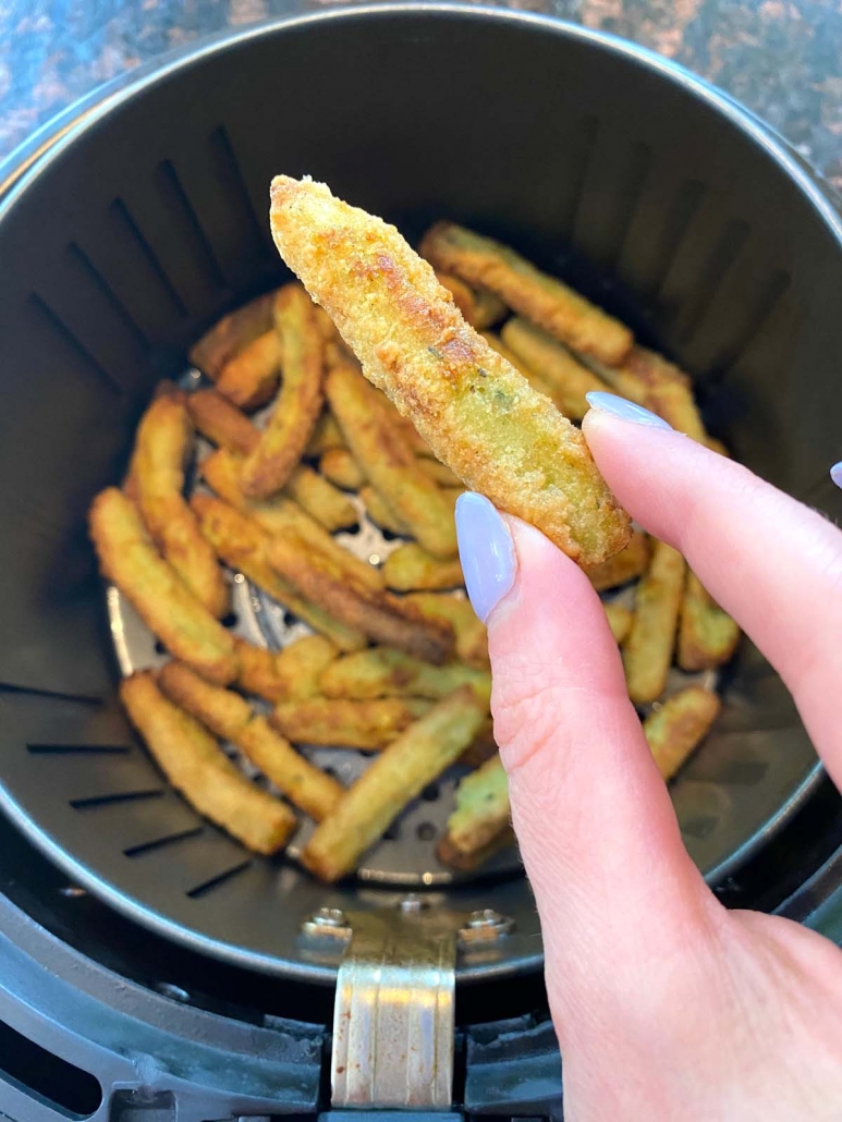 hand holding an air fried zucchini fry above other fries in air fryer