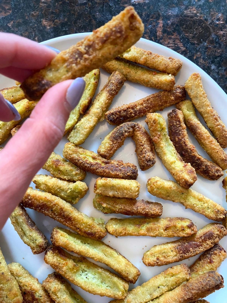 hand holding air fried zucchini fry above plate of other zucchini fries