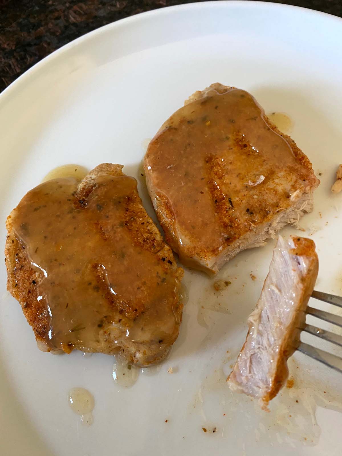 2 pork chops on a plate smothered in gravy with a piece cut out and a fork lifting it up to show juicy inside.