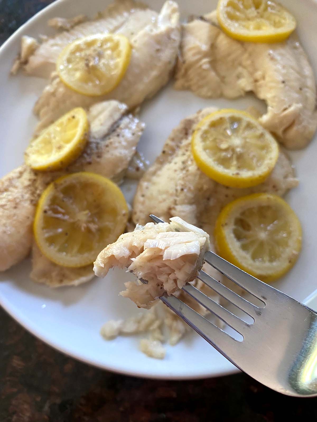 Plate of tilapia with lemon slices and fork showing a piece up close.