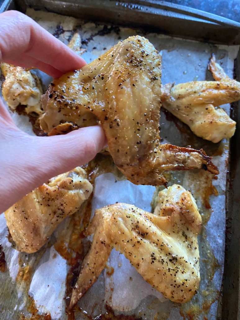 hand holding baked chicken wing in front of baking sheet full of seasoned wings