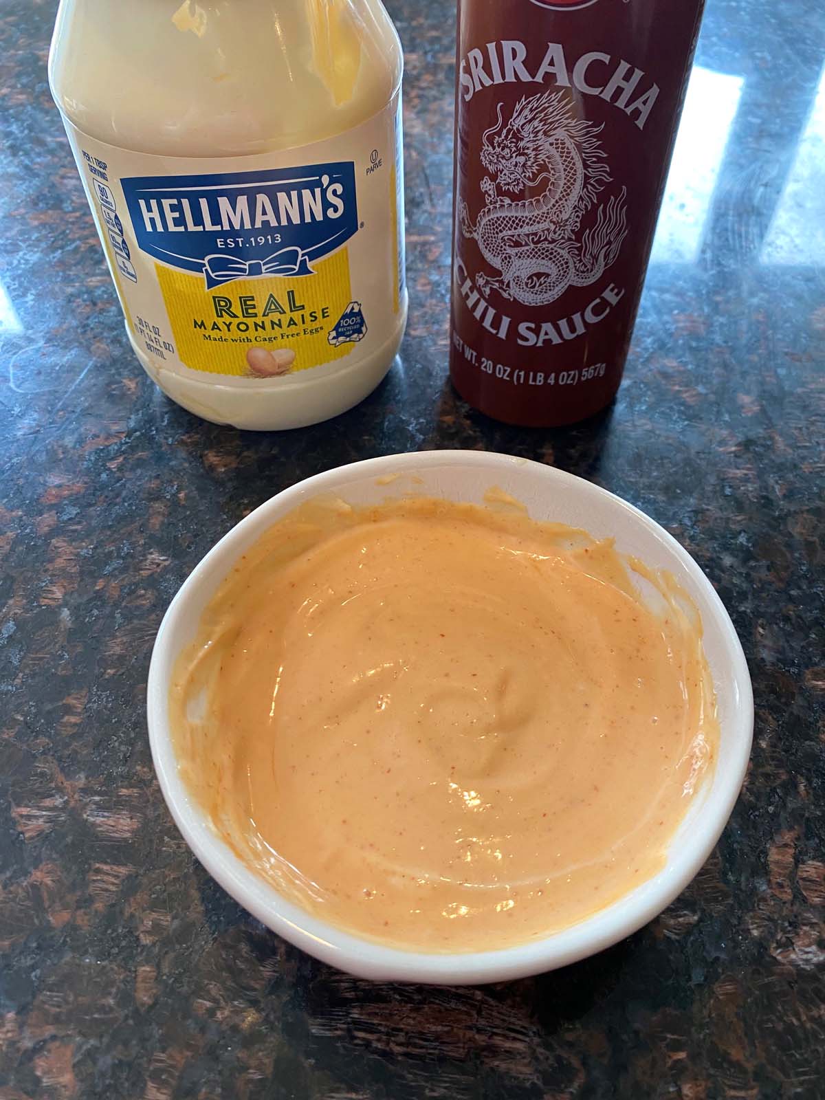 Sriracha spicy mayo in a white bowl with a bottle of sriracha and a jar of mayo.