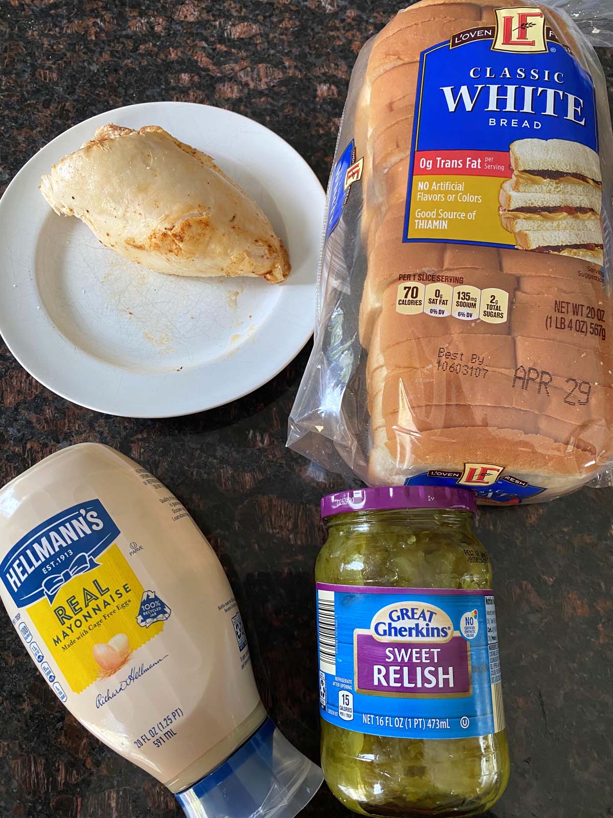 A cooked chicken breast on a plate, tube of mayo, jar of relish, and loaf of bread.