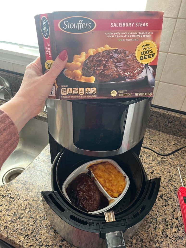 hand holding a package of frozen Salisbury steak dinner in front of an air fryer with a cooked Salisbury steak dinner inside