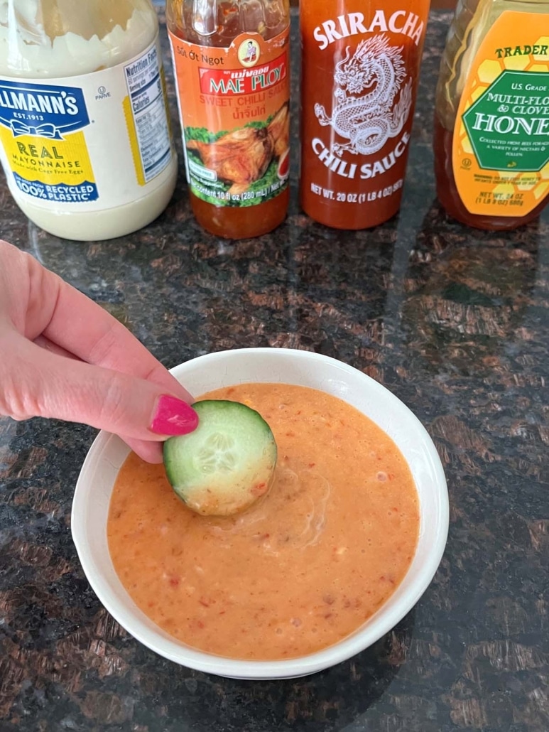 hand dipping a slice of cucumber into bang bang sauce, with mayo, sweet chili sauce, sriracha, and honey behind the bowl