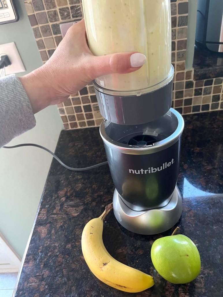 hand placing blender cup on blender, next to an apple and a banana