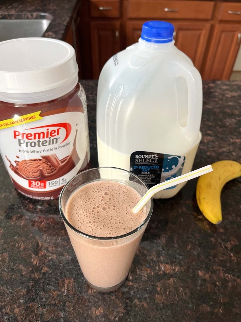 a glass of Chocolate Banana Protein Shake next to a banana, a container of protein powder, and a container of milk