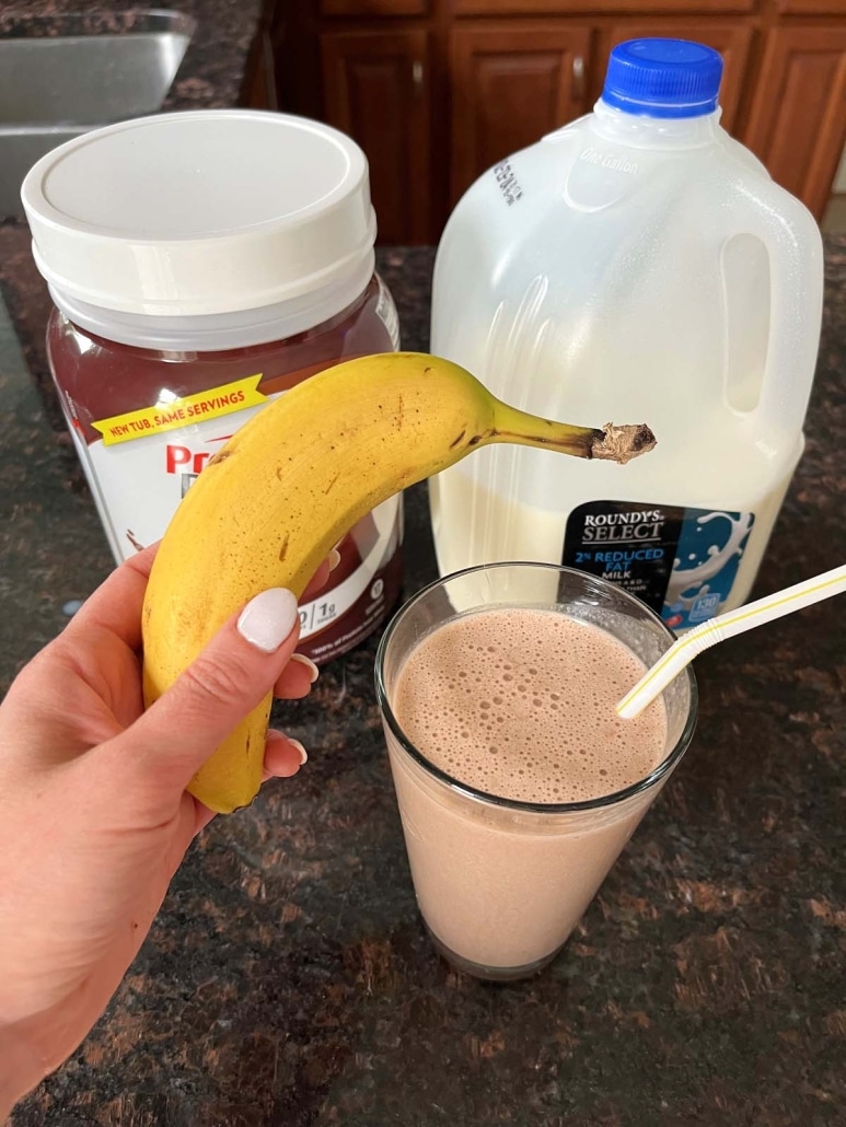 hand holding banana in front of Chocolate Banana Protein Shake, plus chocolate protein powder and milk