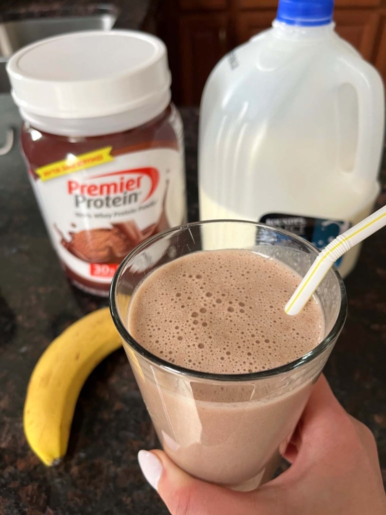 hand holding a Chocolate Banana Protein Shake in front of a banana, canister of protein powder, and a gallon of milk