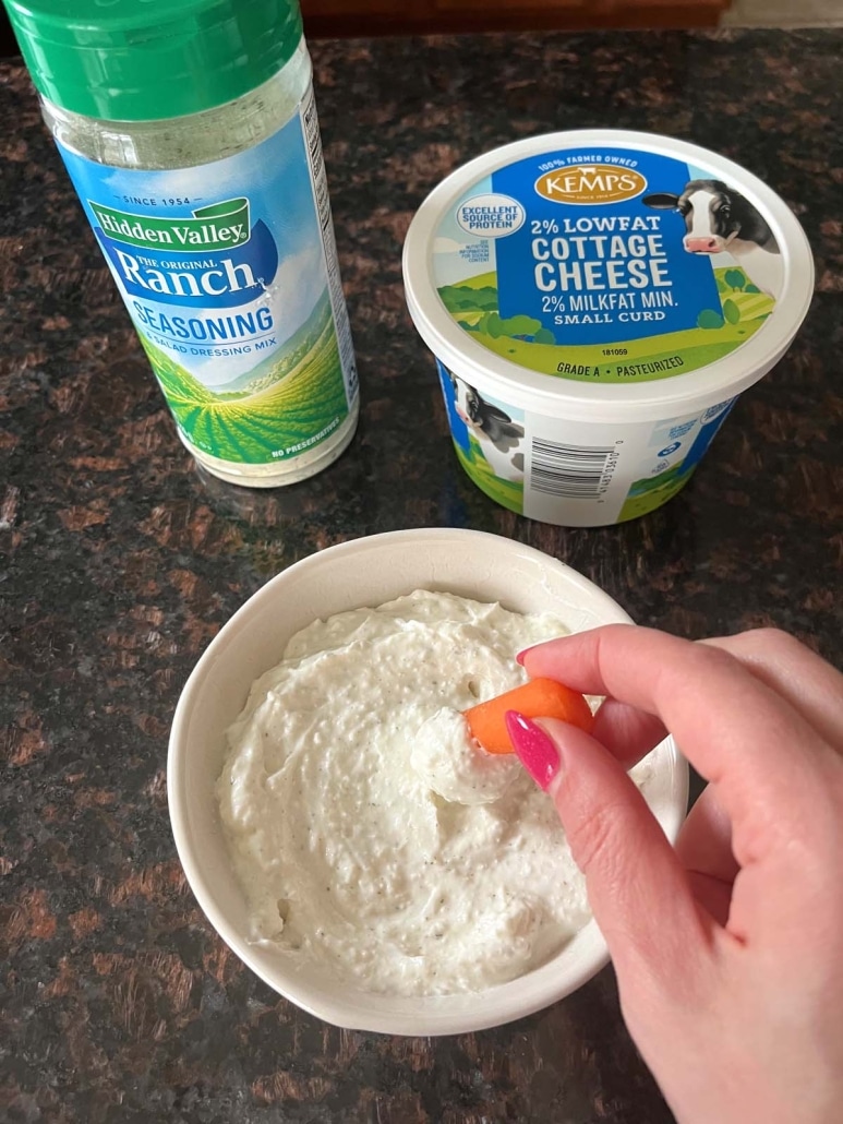 hand dipping a carrot into Cottage Cheese Ranch Dip, next to a container of ranch seasoning and a container of cottage cheese