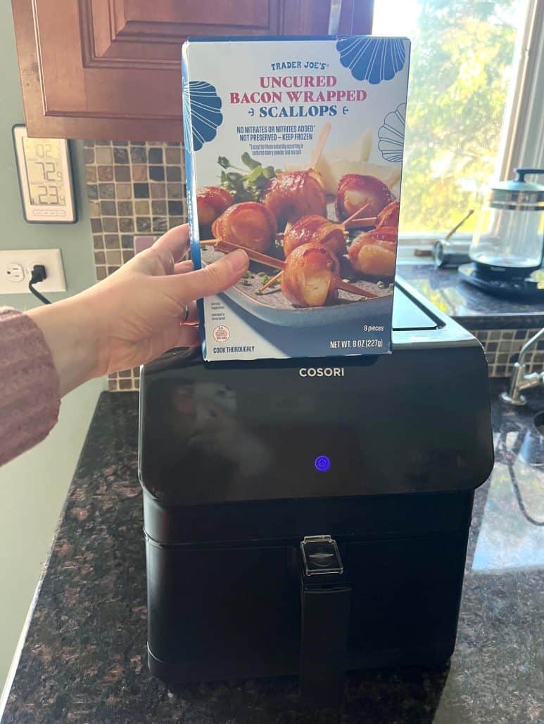 Melanie Mendelson from melaniecooks.com holding a package of uncured bacon-wrapped scallops in front of an air fryer, ready to cook for a quick and crispy seafood appetizer.