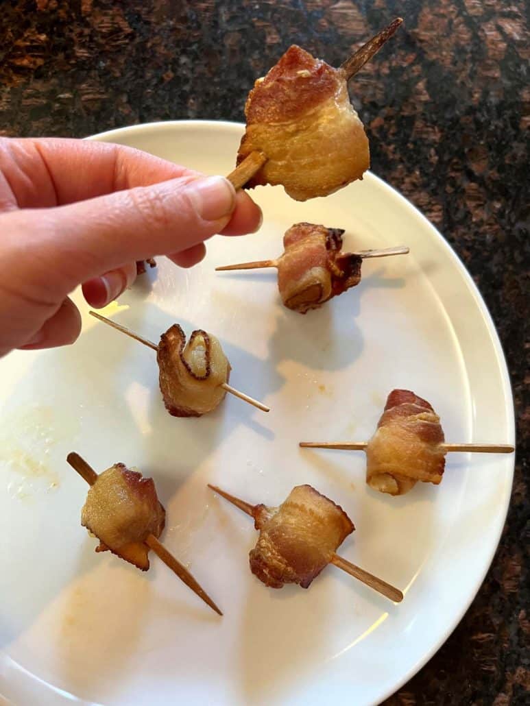 Melanie Mendelson from melaniecooks.com holding a crispy bacon-wrapped scallop on a skewer, with more golden-brown scallops arranged on a white plate in the background.