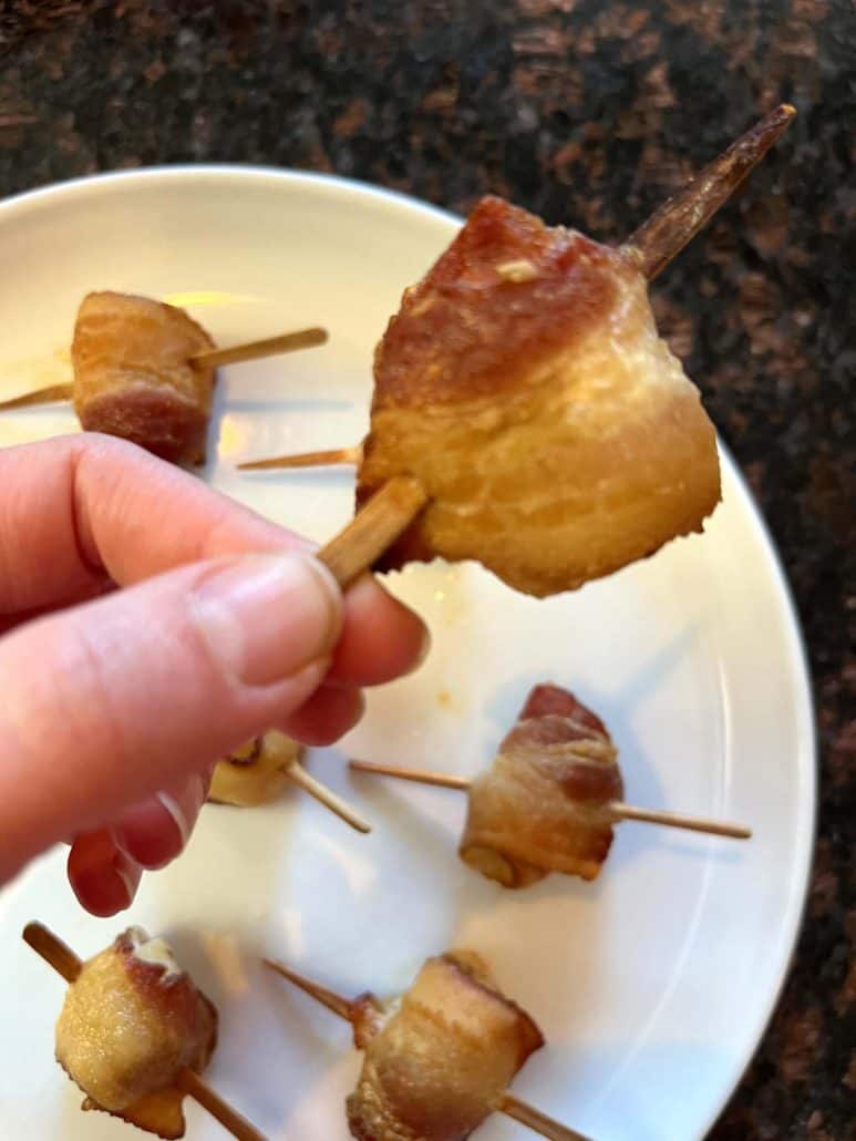 Melanie Mendelson from melaniecooks.com holding a crispy bacon-wrapped scallop on a skewer, with more scallops on a white plate in the background, ready to enjoy.