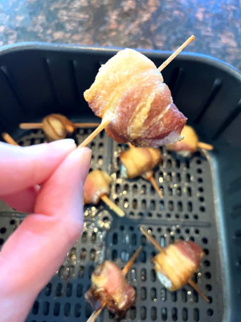 Melanie Mendelson from melaniecooks.com holding a crispy bacon-wrapped scallop on a skewer, fresh from the air fryer, with more golden-brown scallops in the basket below.