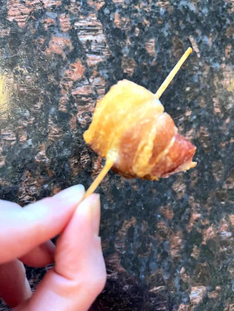 Melanie Mendelson from melaniecooks.com holding a crispy bacon-wrapped scallop on a skewer, highlighting its golden-brown texture and deliciously cooked bacon.