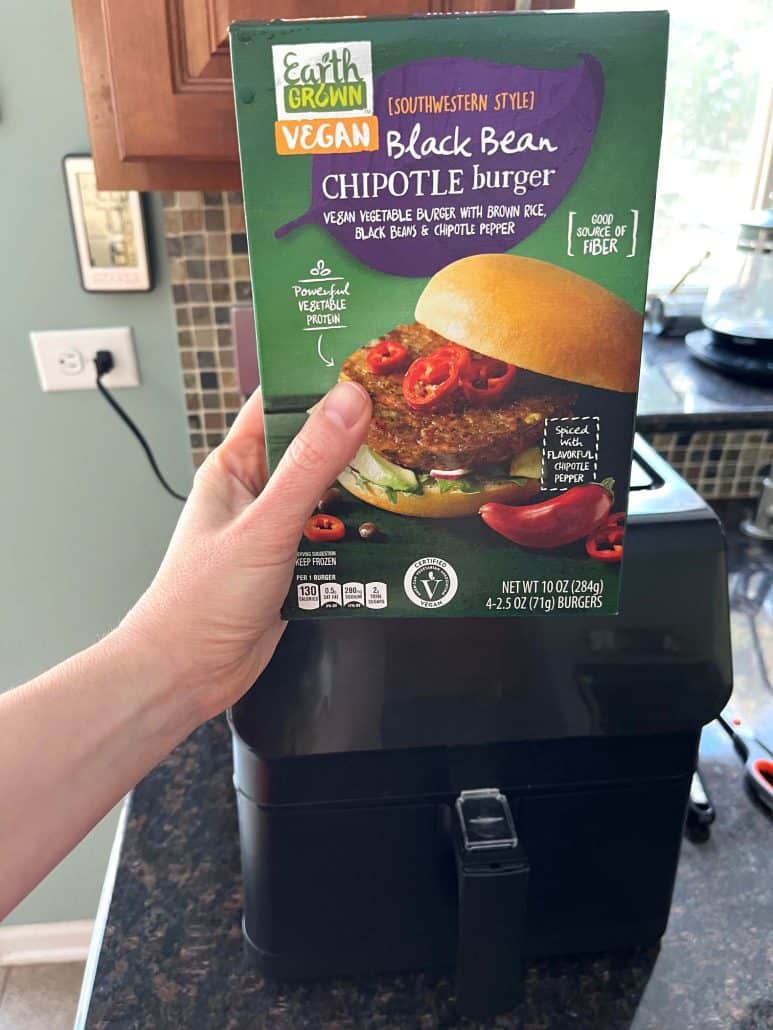 Melanie Mendelson from melaniecooks.com holding a box of frozen black bean chipotle burgers in front of an air fryer, ready to show how to cook them straight from frozen.