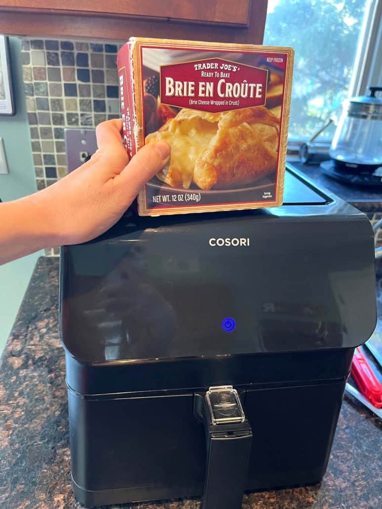 Melanie Mendelson from melaniecooks.com holding a box of Trader Joe’s Brie En Croute in front of the air fryer before cooking.