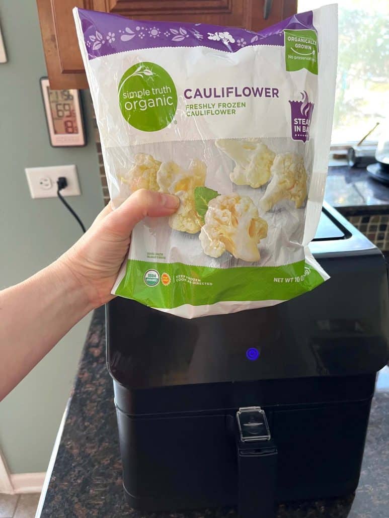 Hand of Melanie Mendelson from melaniecooks.com holding a bag of frozen cauliflower in front of an air fryer, ready to show how to cook.